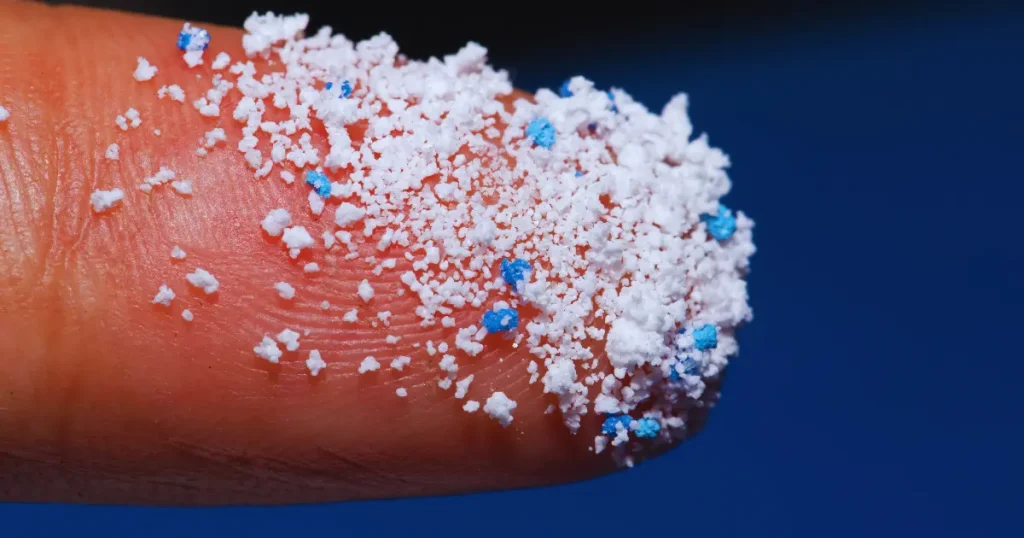Close-up of a fingertip covered in small white and blue microplastic particles, illustrating the scale and texture of microplastic pollution.